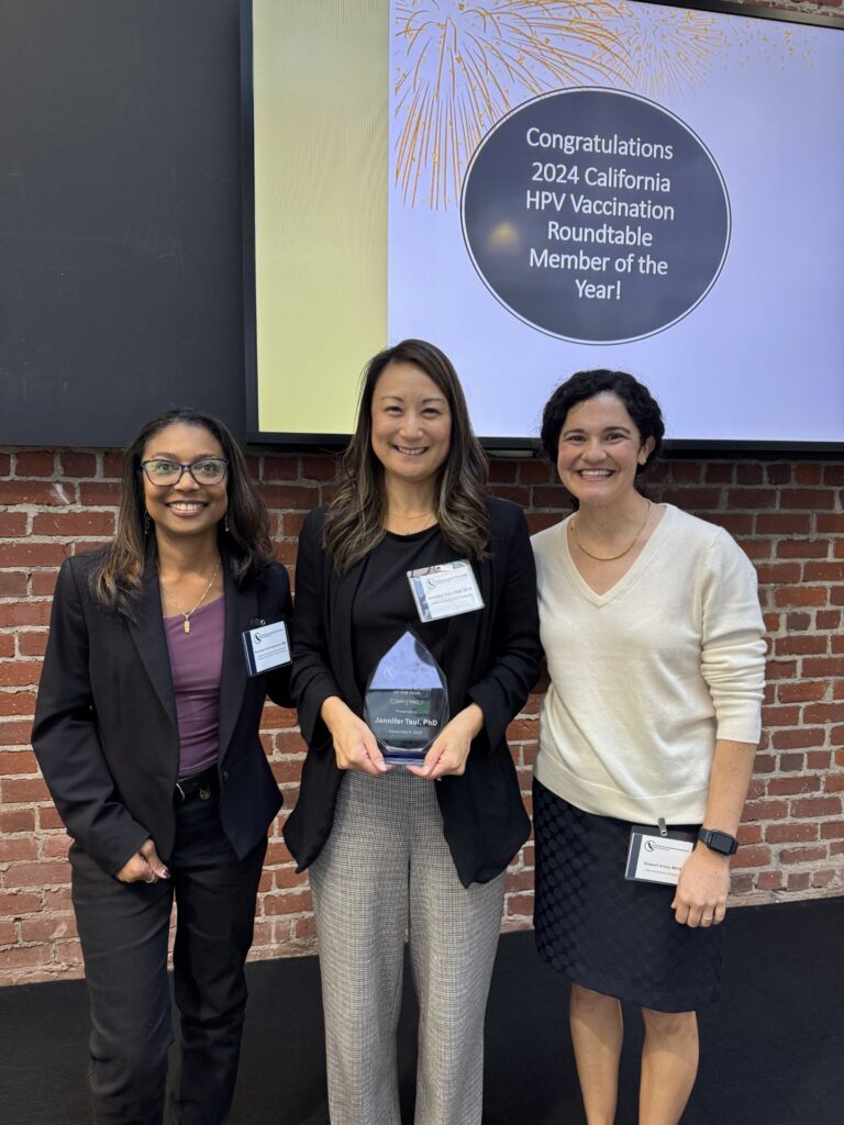 Photo of three individuals standing side by side and smiling. The person in the middle is holding an award.